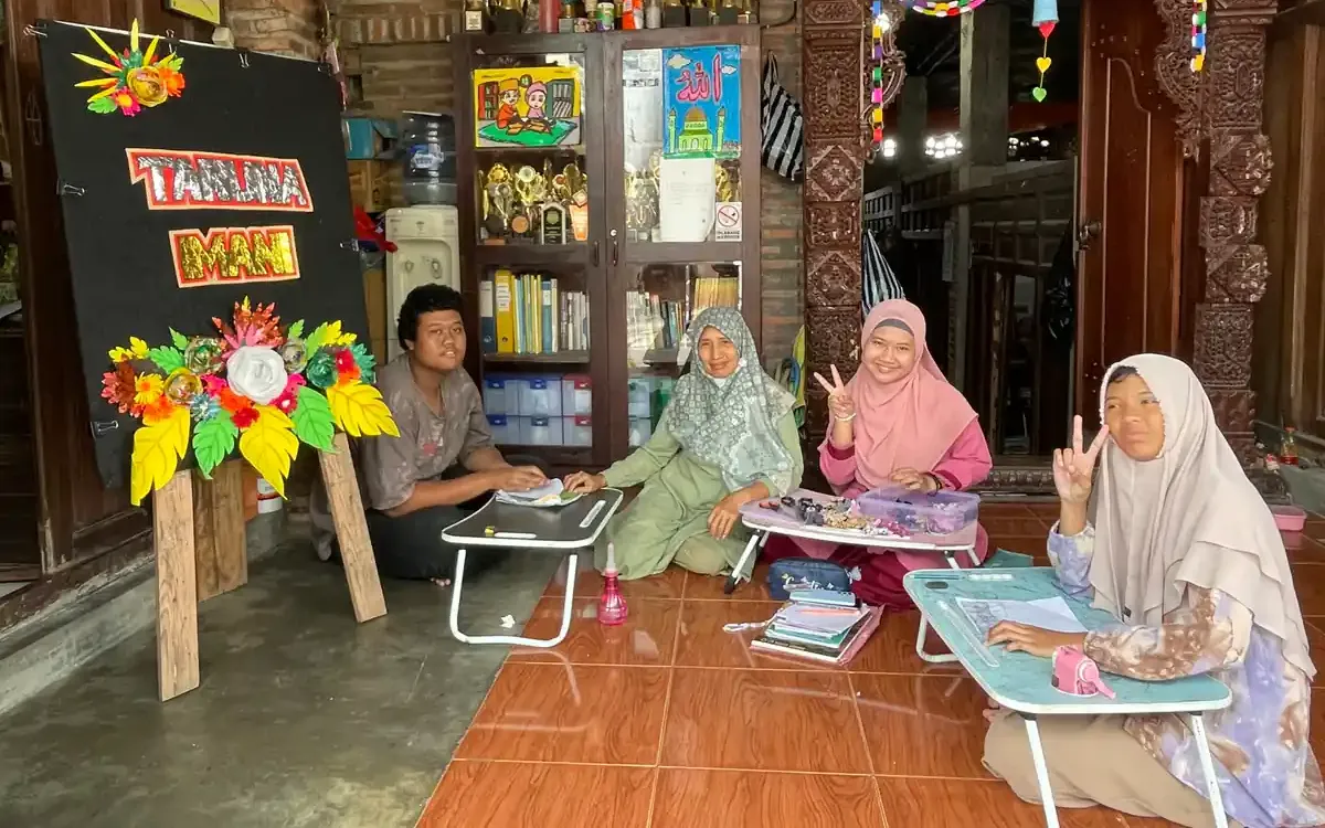 Seorang guru dan beberapa siswa duduk di lantai dengan meja lipat di depan papan bertuliskan Taruna Imani
