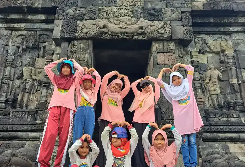 Sekelompok anak berbaju merah muda membentuk tanda hati dengan tangan di depan pintu candi berukir