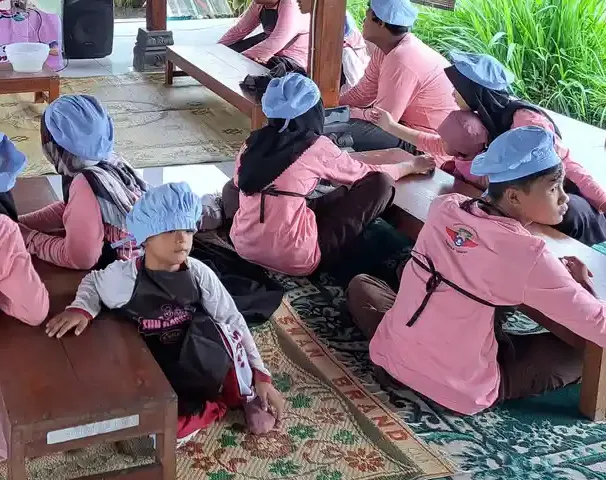 Beberapa anak bertopi kain biru dan bercelemek duduk menghadap meja kayu rendah di sebuah pendopo