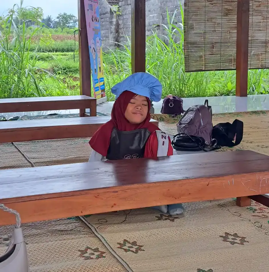 Seorang anak bertopi koki dan bercelemek duduk di bangku kayu panjang di pendopo, tas peserta tertata di belakangnya