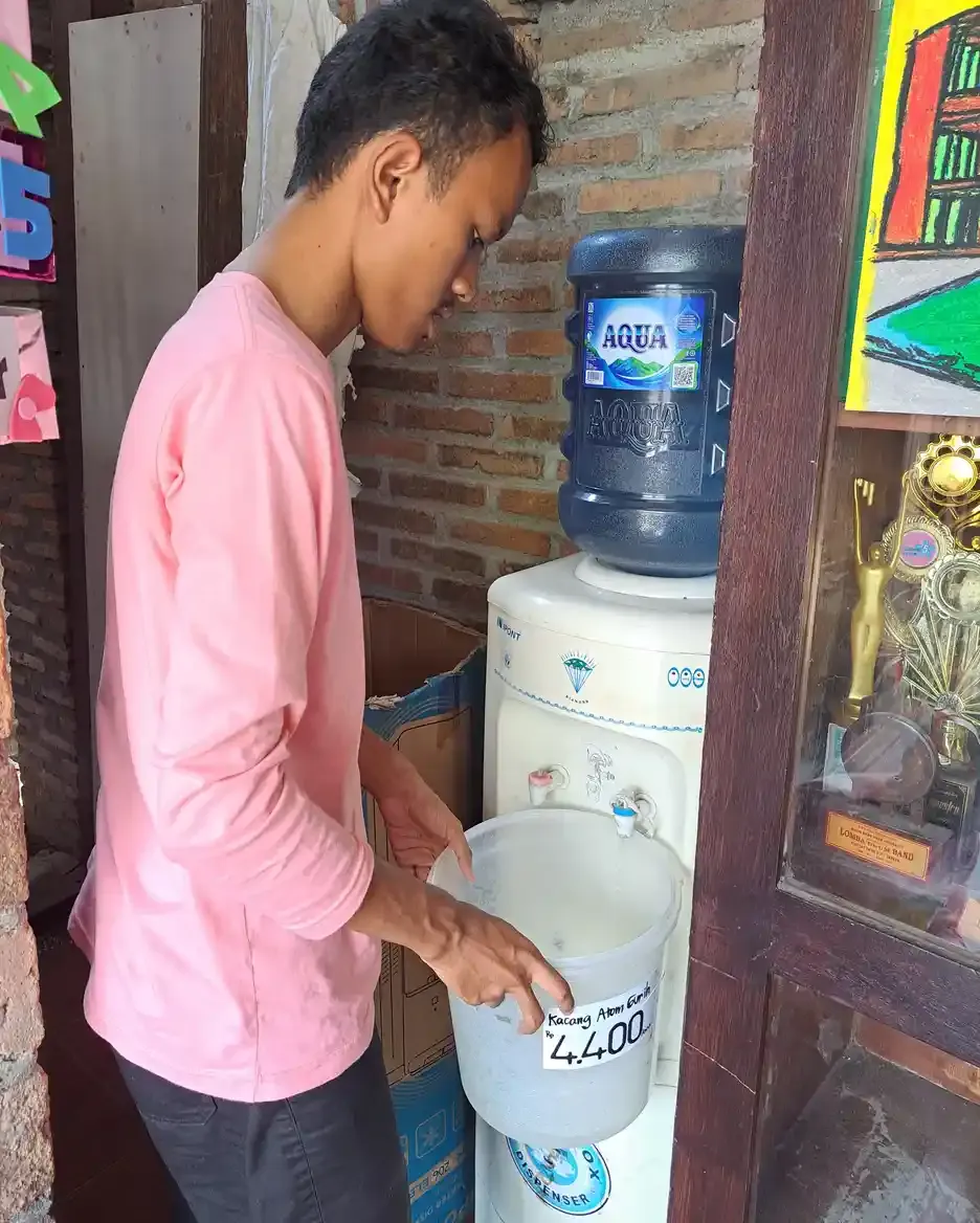 Seorang remaja berkaus merah muda mengisi wadah plastik dari dispenser air minum di sudut ruangan berdinding bata