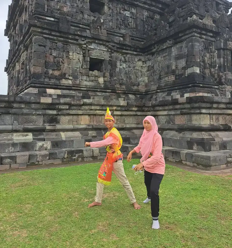 Seorang siswa berkostum tari dan seorang pendamping berkerudung merah muda bergerak bersama di halaman rumput depan candi batu