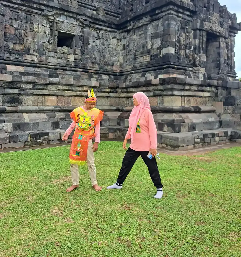 Seorang siswa berkostum tari tradisional dan seorang pendamping berkerudung merah muda bergerak bersama di halaman rumput depan candi