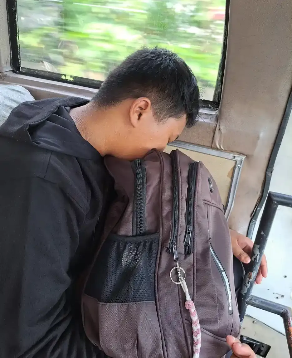 Seorang remaja berjaket hitam memeluk tas punggungnya sambil beristirahat di bangku dekat jendela kendaraan