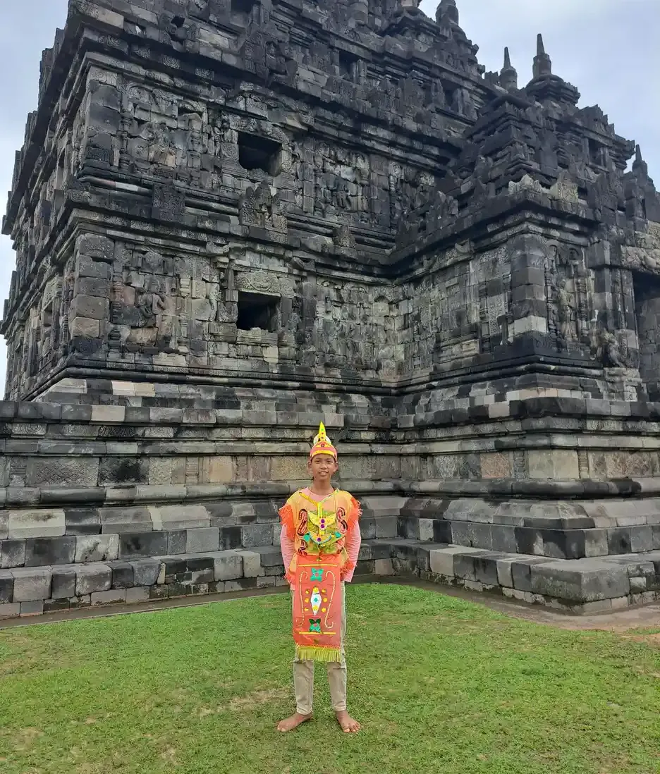 Seorang siswa mengenakan kostum tari tradisional berwarna oranye berdiri di halaman rumput di depan candi batu
