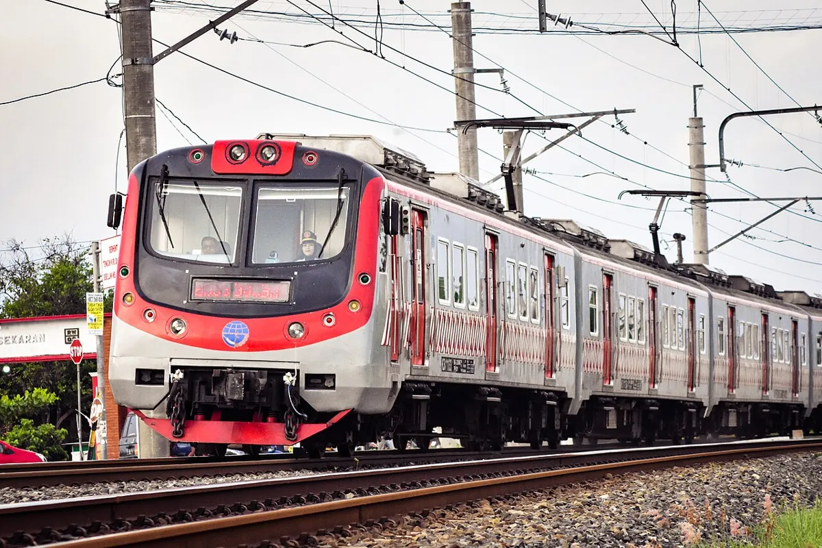 Rangkaian KRL berwarna merah dan putih melaju di atas rel di bawah kabel listrik aliran atas