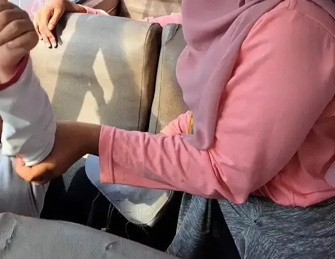 Pendamping berbaju merah muda memegang lengan peserta berbaju putih yang duduk di kursi berlapis kulit