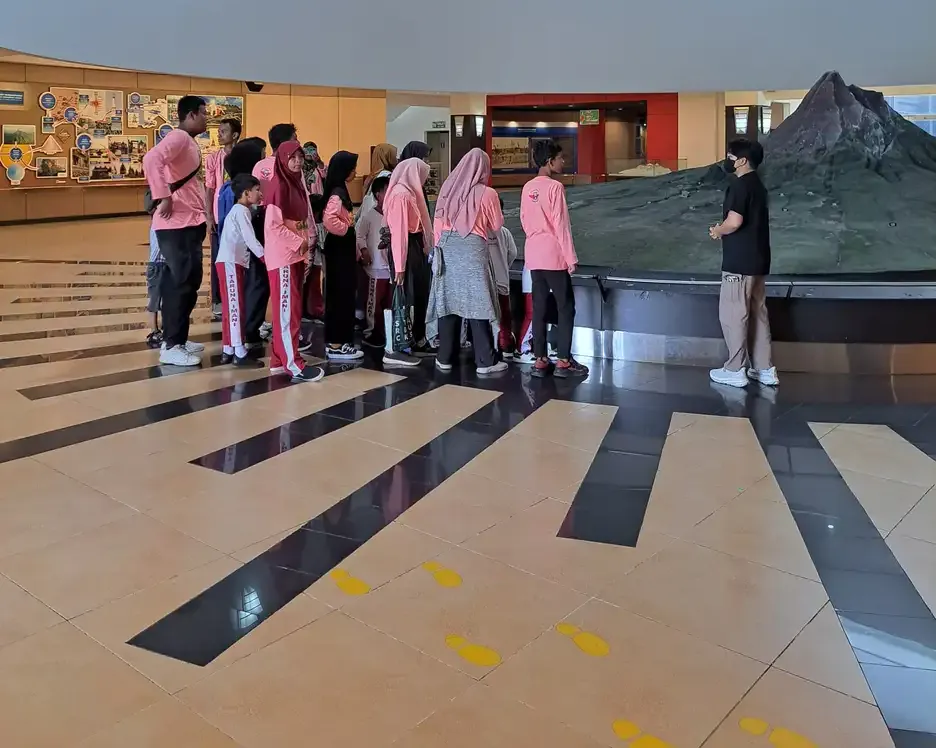 Rombongan anak berseragam dan pendamping berbaris di aula museum yang luas dengan lantai bergaris dan maket gunung
