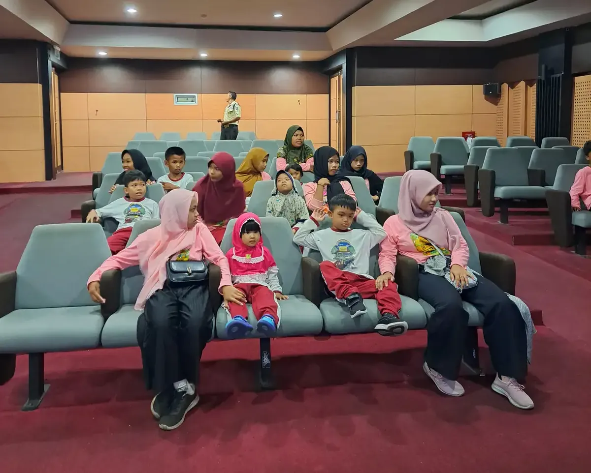 Anak-anak dan pendamping duduk berjajar di kursi auditorium berkarpet merah, sebagian menoleh ke arah kamera