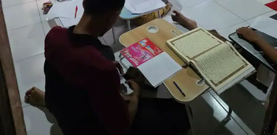 Seorang peserta duduk menghadap meja lipat berisi mushaf Al-Quran terbuka dan buku catatan, difoto dari belakang