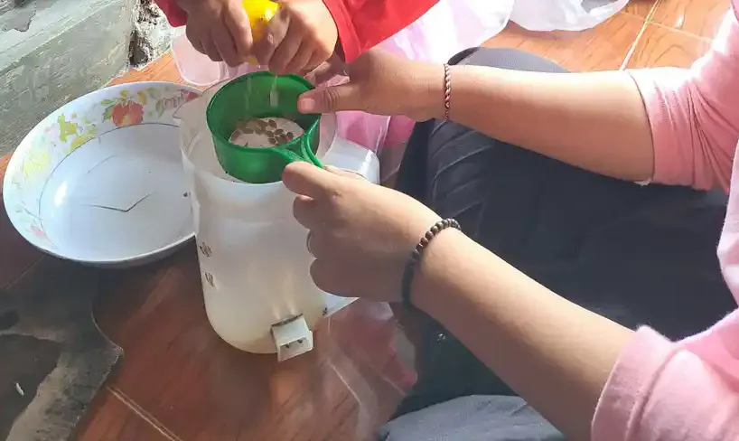 Tangan anak dan tangan pendamping bersama-sama memegang corong hijau di atas wadah plastik