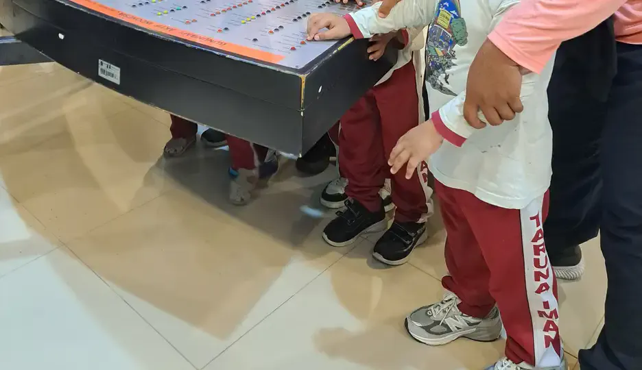 Beberapa anak berseragam berdiri di sekeliling meja permainan interaktif di lantai keramik museum, tangan mereka menyentuh permukaan meja