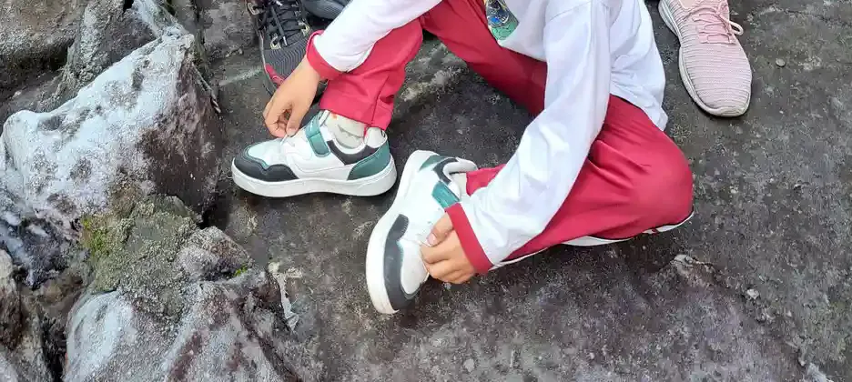 Seorang anak duduk di atas batu besar sambil memegang sepatu kets putihnya dengan kedua tangan