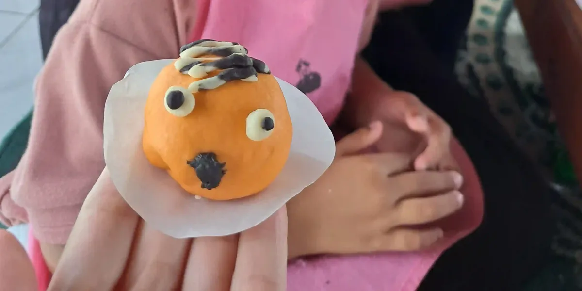 Tangan orang dewasa menyodorkan kue bulat oranye berhias wajah lucu, dengan tangan anak berbaju merah muda di latar belakang