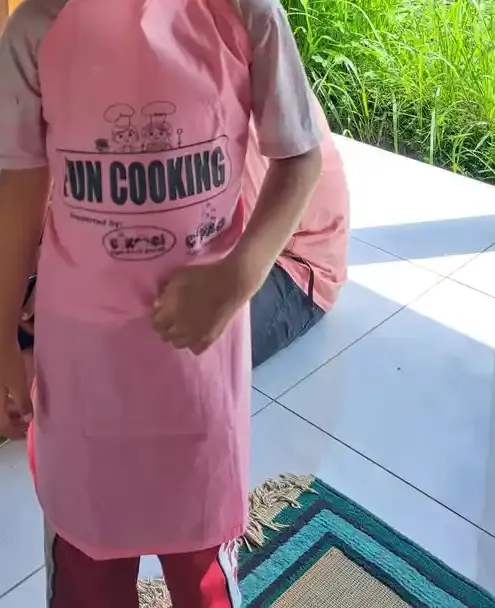 Seorang anak bercelemek merah muda bertuliskan Fun Cooking berdiri di lantai keramik paviliun