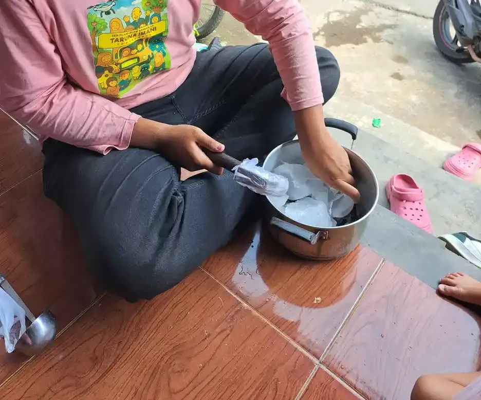 Seorang anak berkaus merah muda duduk bersila di lantai teras sambil mengaduk isi panci dengan spatula