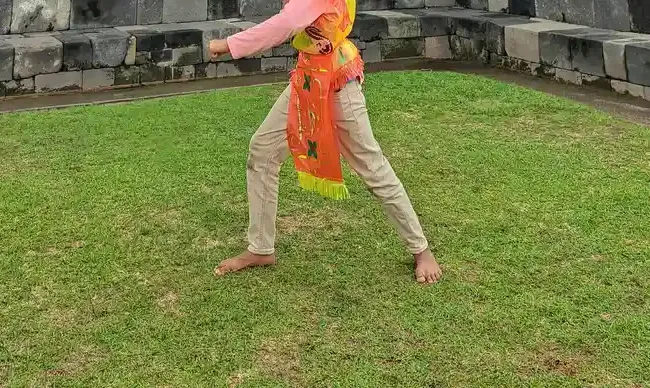 Kaki seorang anak bercelana krem dan berselendang oranye berdiri melebar di atas rumput dengan dinding batu candi di belakangnya