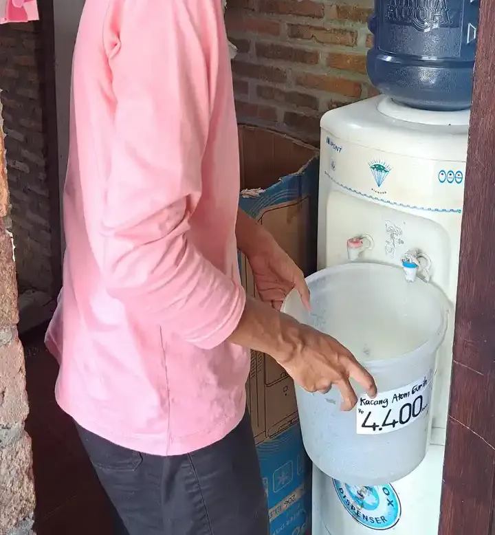 Seseorang berkaus merah muda memegang ember plastik di depan dispenser air galon